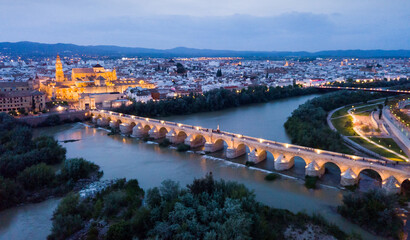 Scenic aerial view of lighted Roman bridge across Guadalquivir river and Mezquita-Catedral on background with Cordoba cityscape at twilight, Spain.. - obrazy, fototapety, plakaty