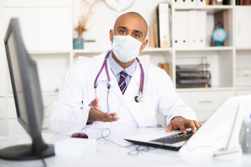 Doctor in protective medical mask sitting at workplace with computer in her office
