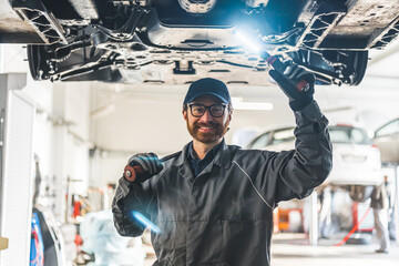 Smiling auto mechanic with work light standing under the lifted car . High quality photo