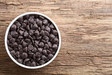Chocolate chips or morsels in white bowl, photographed overhead on wood with copy space on the side (Selective Focus, Focus on the top chocolate chips)