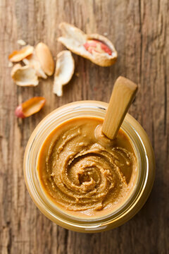 Creamy Smooth Peanut Butter In Jar With Wooden Spoon, Peanuts In Shell Around, Photographed Overhead On Wood (Selective Focus, Focus On The Peanut Butter)