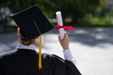 Fototapeta Rear view of a caucasian woman in a graduate gown holding a diploma outdoors.  obraz
