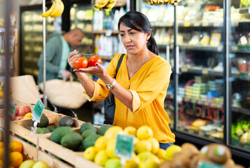 Fototapeta Hispanic female shopper looking for fresh organic tomatoes in greengrocery department of supermarket obraz