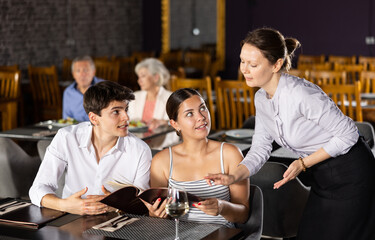 Fototapeta Adult female waiter in uniform takes order from young couple in restaurant obraz