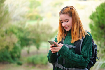 Happy redhead young female hiker consulting smartphone while trekking.