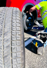 Flat tire concept, a piece of metal pricks into car tire while driving. Blur background of a technician fixing by replacing a spare tire on the road. Close up unused tyre with defocus of a repair man.