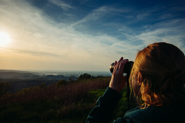 A hiker girl looks through binoculars at nature and birds standing on the top of a mountain in the bright rays of setting sun against the background of beautiful spring nature and cloudy blue sky