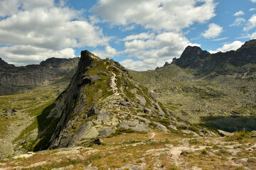 A narrow hiking trail going up the steep slope of a high mountain overlooking picturesque cliffs on a cloudy summer day.