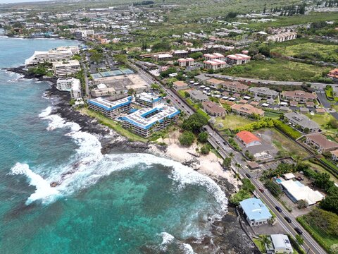 View of coastal Kailua-Kona on the Big Island of Hawai'i. Aerial drone looking along the coast with palm trees, Honl’s Beach, condominiums, coastal highway, and blue and green waters.