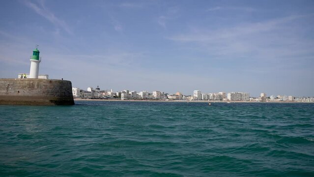 Green top lighthouse at the port town of Sables D'olonne in Western France with people beside it, Pan left shot from boat