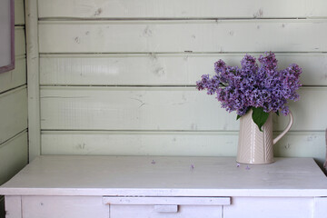 A bouquet of lilacs in a jug on a white table. Country house interior with wooden walls.