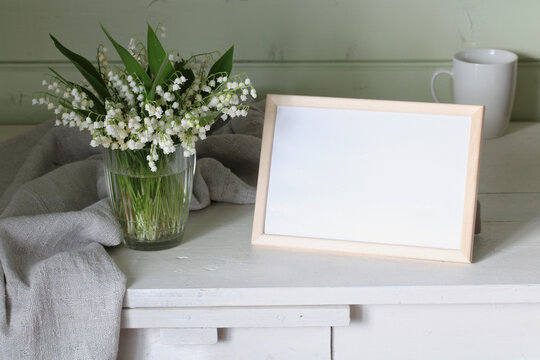 An empty photo frame on the table. A bouquet of forest lilies of the valley in a glass mug. Copy space.