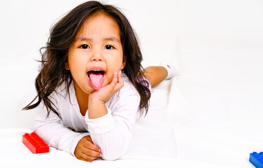 Stylish Asian 3-year-old girl lies and shows her tongue, loose hair, posing on a white background. for advertising with copy space