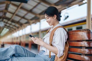 Asian teenage female traveler using her smart phone mobile while waiting for a train at a station.