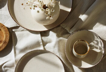 Tableware on a textile napkins, sunlight shadows top view with copy space.Modern kitchen tableware and vase with flowers.Neutral beige color.Business food brand template