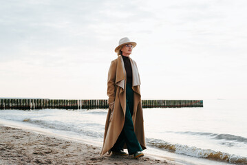 Thoughtful stylish mature woman walking on beach by sea. Alone senior woman in brown coat on seashore outdoors