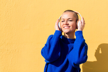 woman with a shaved head and a blue jumper enjoying happy the music in her headphones with a yellow wall in the background, concept of rhythm and positive people, copy space for text