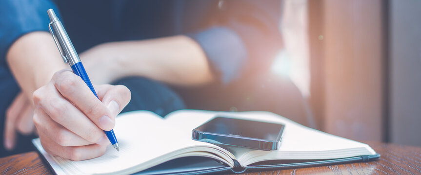 Woman hand writing on a notepad with a blue pen in the office.There is a mobile phone on the table.