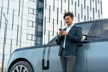 Young businessman standing near luxury car and using smartphone