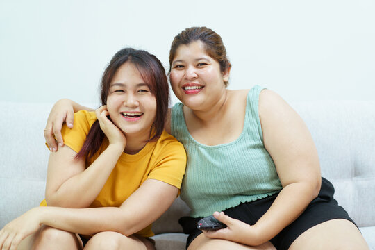Happy cheerful Asian women sitting on a sofa in living room and watching a movie on tv together, big size fat women friends or LGBTQ+ lesbian couple relaxing in living room and watching television.