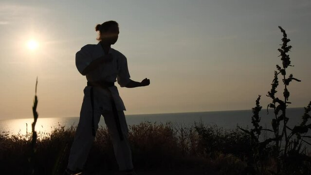 Karate training "shadow boxing" in the backlight of the dawn sun against the backdrop of the sea. The girl makes blows to an imaginary opponent. Eastern martial arts.