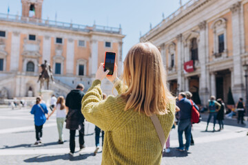 Blonde young Woman with smartphone is walking on a sunny day. Capitol in Rome, Piazza del Campidoglio in Capitoline Hill, Italy. Concept of traveling famous landmarks.