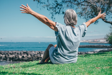 Fototapeta na wymiar Back view of senior woman sitting barefoot in meadow in front of sea looking at the horizon with open arms, relaxed senior lady enjoying free time on vacation or retirement