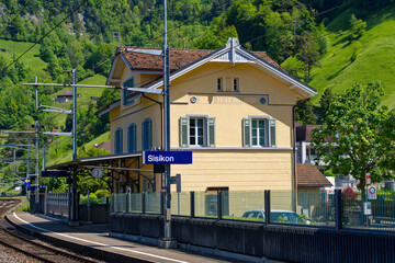 Blue and white sign with yellow railroad building at platform of railway station at village of Sisikon on a sunny spring day. Photo taken May 22nd, 2023, Sisikon, Switzerland.