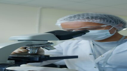 Vertical medium closeup of Biracial female scientist wearing mask, goggles and hairnet adjusting and looking through microscope in laboratory at daytime - Powered by Adobe