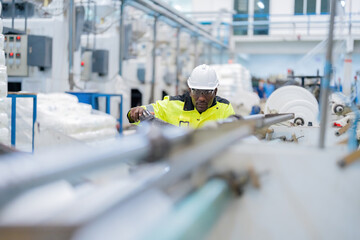 African male engineer is a mechanic He is sitting attentively and determinedly inspecting machines that are working electronically, wearing a vest and helmet in a steel and plastic industry.