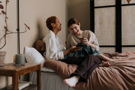 Happy non-binary couple sharing smart phone while sitting on bed at home - Powered by Adobe