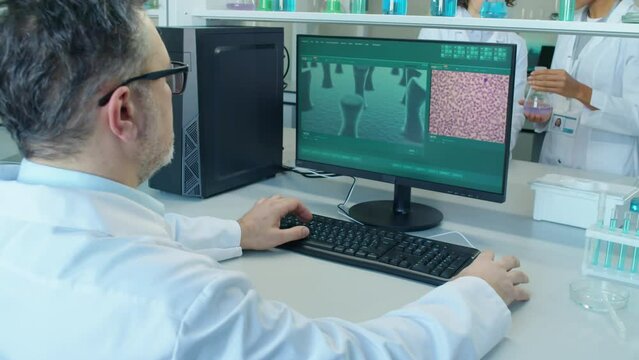 High angle focus on male Caucasian professor sitting at desk in laboratory typing something on keyboard looking at computer screen with cells and molecules on