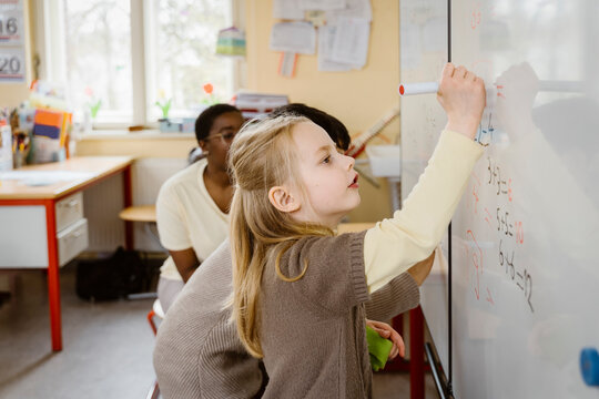Blond girl solving maths problem while writing on whiteboard in classroom