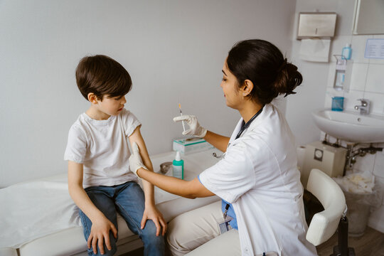 Side View Of Female Pediatrician Giving Vaccine To Boy Sitting On Bed In Clinic At Hospital