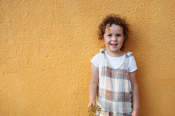 Happy cute kid child girl with curly hair, girl in stylish overalls holding a plant, orange color background with copy space