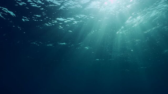 View of sun light from blue abyss, slow motion. Light filters down through blue water. Underwater sun rays in depth ocean. Underwater sun light shine under deep water with ripples on waves surface