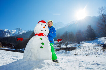 Obraz premium Young boy in winter outfit stand with freshly build snowman