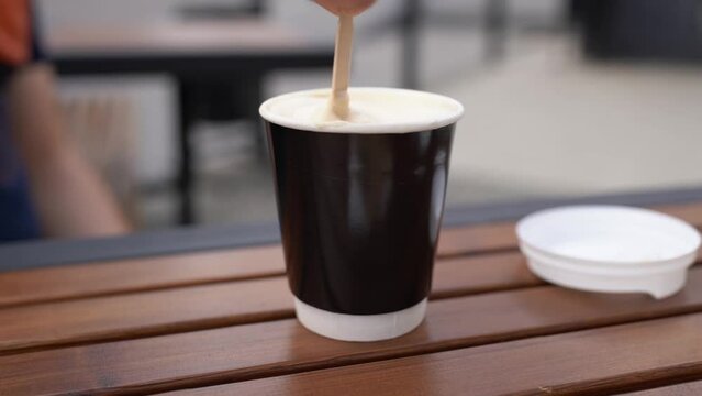 Slow and fast stirring sugar in a glass in a street cafe. Hand holding a wooden spoon and quickly stir cappuccino with white milk foam in a black paper cup outdoor. Urban coffee to go footage concept