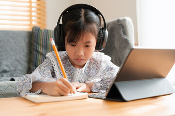 Asian little girl doing homework, using digital tablet with headphone.  - 610574364