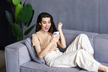Woman with phone and celebration on sofa, winning and smile with success, profit or online gambling...