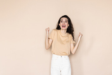 Young woman posing doing winner gesture celebrate clench fists say yes isolated on beige background studio.