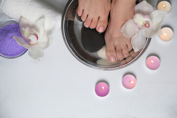 Woman soaking her feet in bowl with water, flower and spa stones on white marble floor, top view. Pedicure procedure