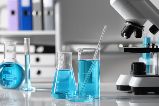Different laboratory glassware with light blue liquid near microscope on table