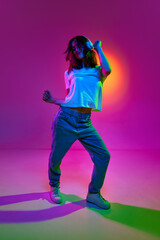 Portrait of a young girl wearing jeans, white t-shirt and headphones listening to music, dancing with pleasure over pink background in neon light