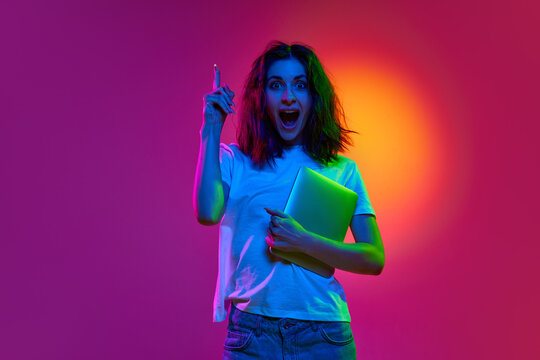 Portrait of pensive, young student, girl holding tablet showing finger with open mouth over pink studio background in neon light. Concept of brainstorming, insight