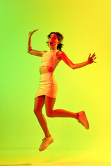 Funny portrait of emotive, young girl wearing skirt and top jumping up over acid green color studio...
