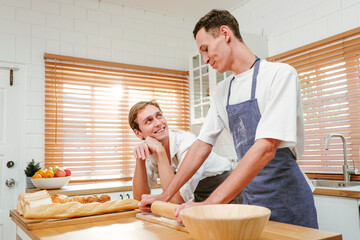 Fototapeta premium Happy Caucasian gay couple cooking together in the kitchen while watching each other with fun and a smile. Man standing by boyfriend preparing food at home. LGBT couple concept.