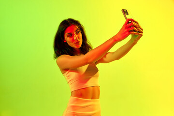 Fototapeta Portrait of brunette girl, woman with wavy hair taking selfie by smartphone over gradient studio background in neon light. Beauty, youth, human emotions obraz