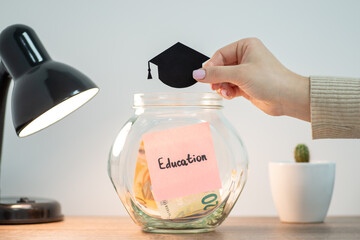 Fototapeta Girl holds picture of black student`s hat above the glass jar with some money and sticker with word Education on it, close up. Concept of investment into future profession and knowledge obraz