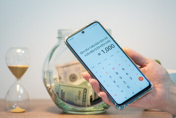 Fototapeta Mobile phone with calculation on its screen, glass jar with dollar banknotes and sandglass on the table on the background. Money calculation or financial investment, savings planning concepts obraz